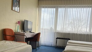 A hotel room with two beds, a desk, a chair, a television, and a window with sheer curtains. - AM Flughafen (KOLN)