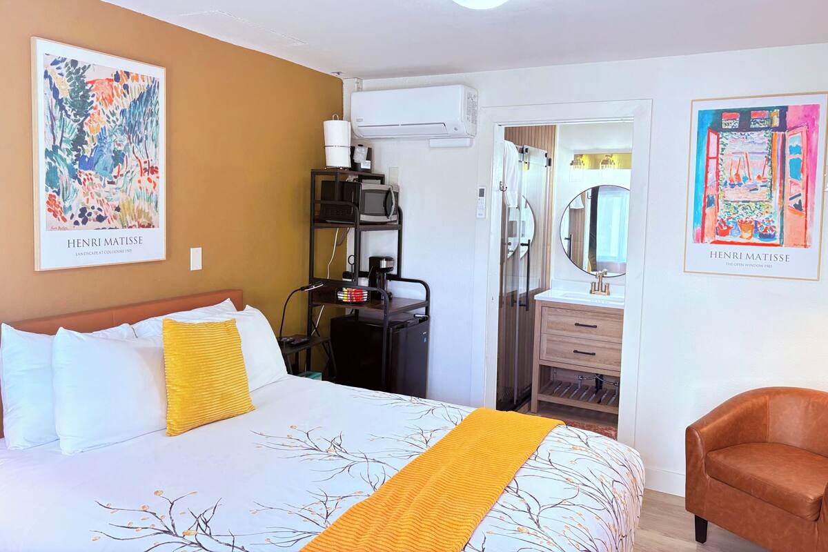 A hotel room with a bed, a desk with a computer, an air conditioner, a bathroom with a mirror, and two Henri Matisse paintings.