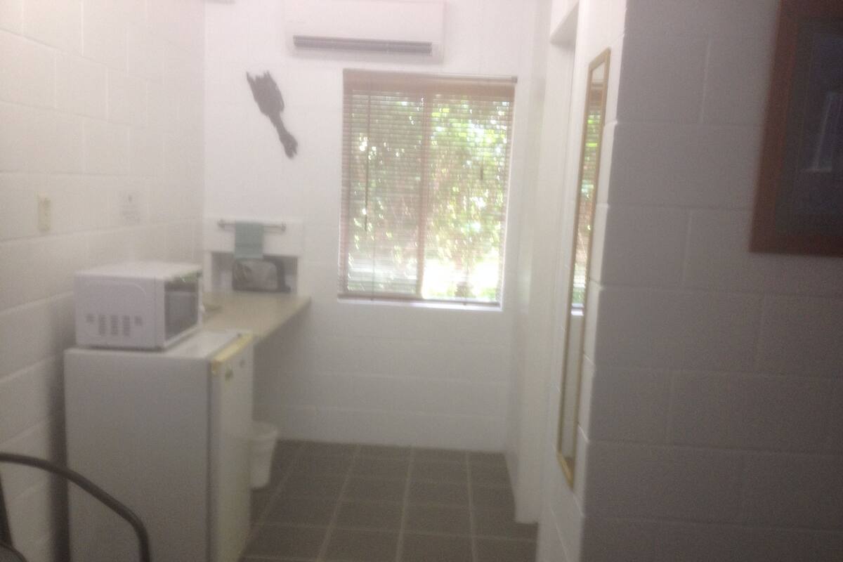 Una cocina pequeña y poco iluminada, con un microondas y un lavabo. Hay una ventana con persianas y un marco de foto en la pared.