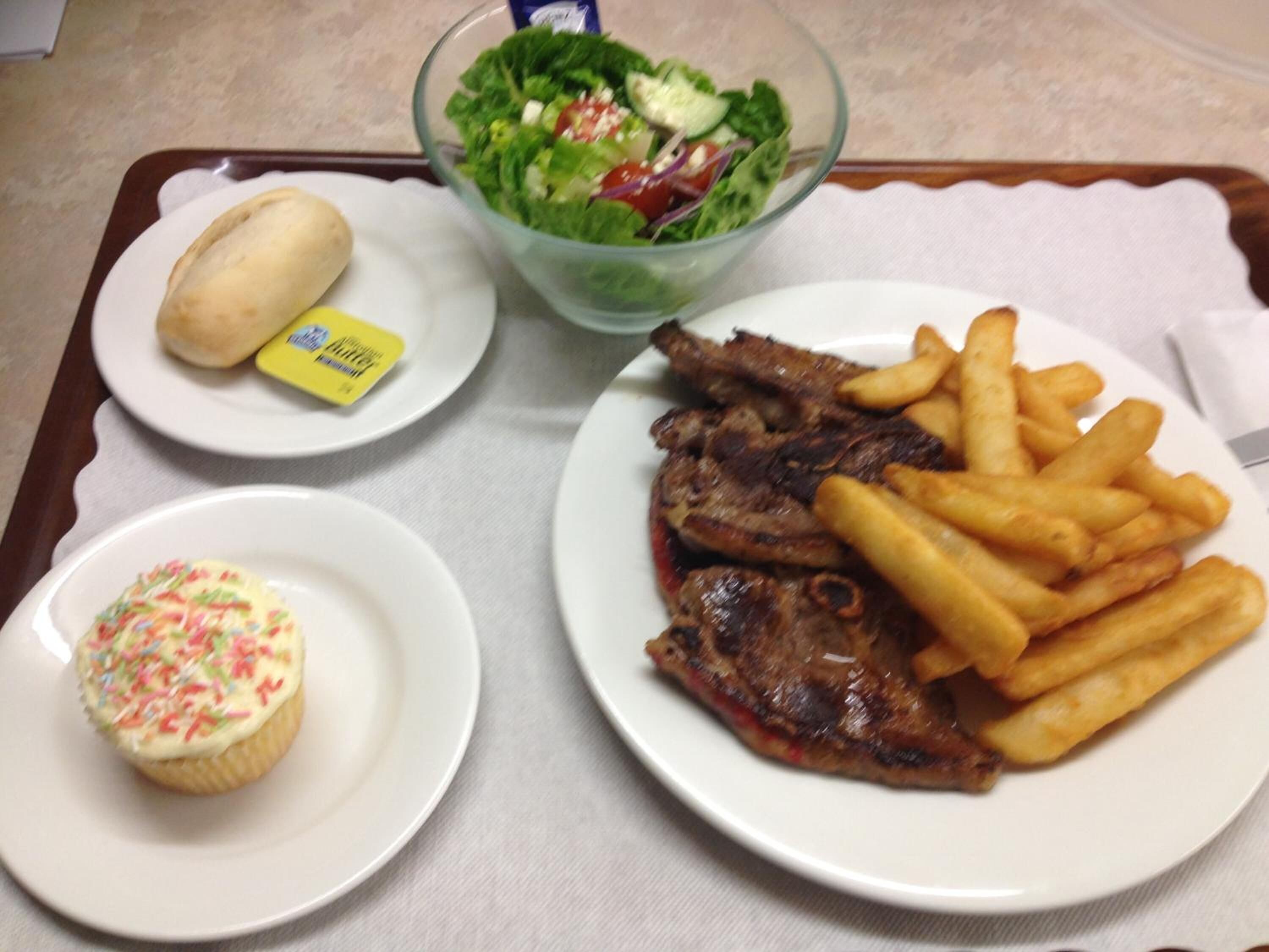A tray with a plate of steak and fries, a cupcake, a salad, and a bun with butter.