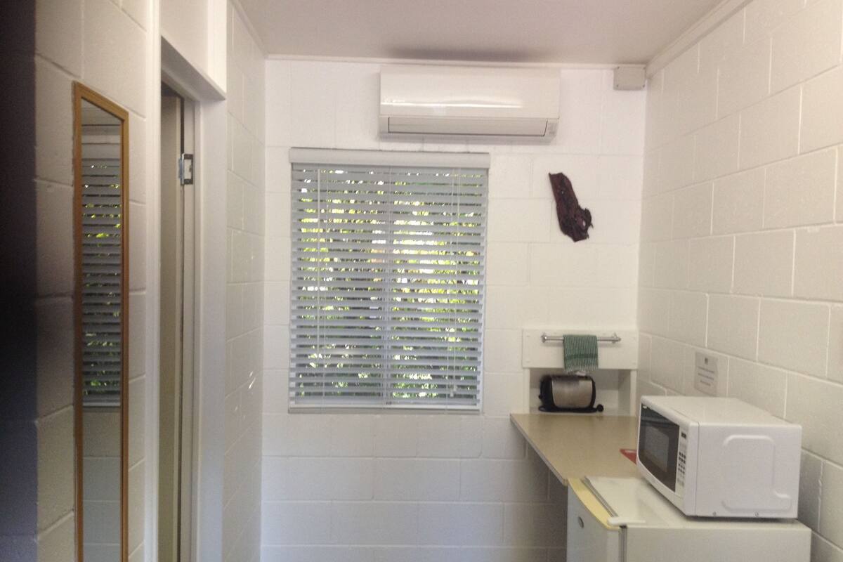 Una pequeña kitchenette con microondas, una ventana con persianas y una toalla colgada en un perchero.