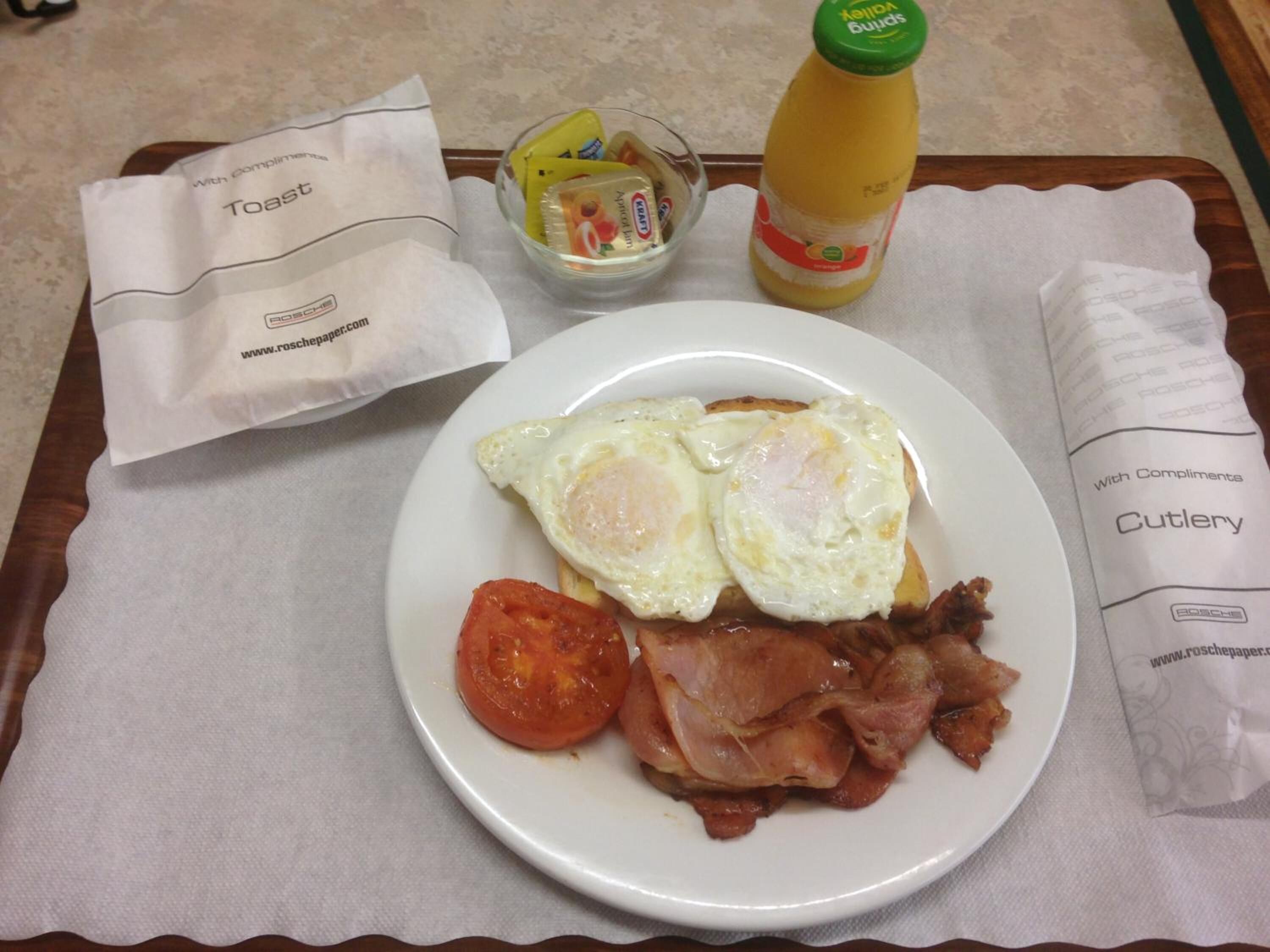 Ein Frühstücksbrett mit Toast, Eiern, Speck und einer Tomate, sowie Gewürzen und einer Saftflasche.