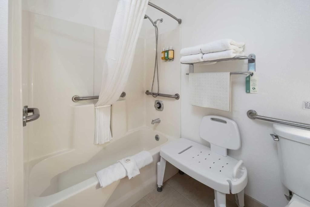 A bathroom with a shower, a toilet, a grab bar, and a white bench.