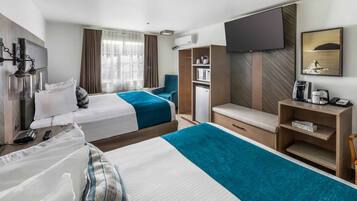 A hotel room with two beds, a TV, a desk with a coffee maker, a mini fridge, and a view of the sea.