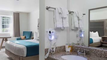 A hotel room with a bed, a sitting area with a chair, a large window, a bathroom with a sink, a mirror, and two hair dryers.