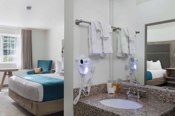 A hotel room with a bed, a sitting area with a chair, a large window, a bathroom with a sink, a mirror, and two hair dryers.