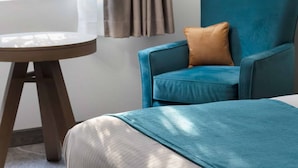 A room with a bed, a blue armchair with a brown pillow, a round wooden table, and a window with curtains. - The Marina Inn (Anacortes)