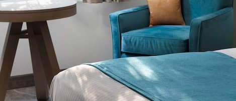 A room with a bed, a blue armchair with a brown pillow, a round wooden table, and a window with curtains.