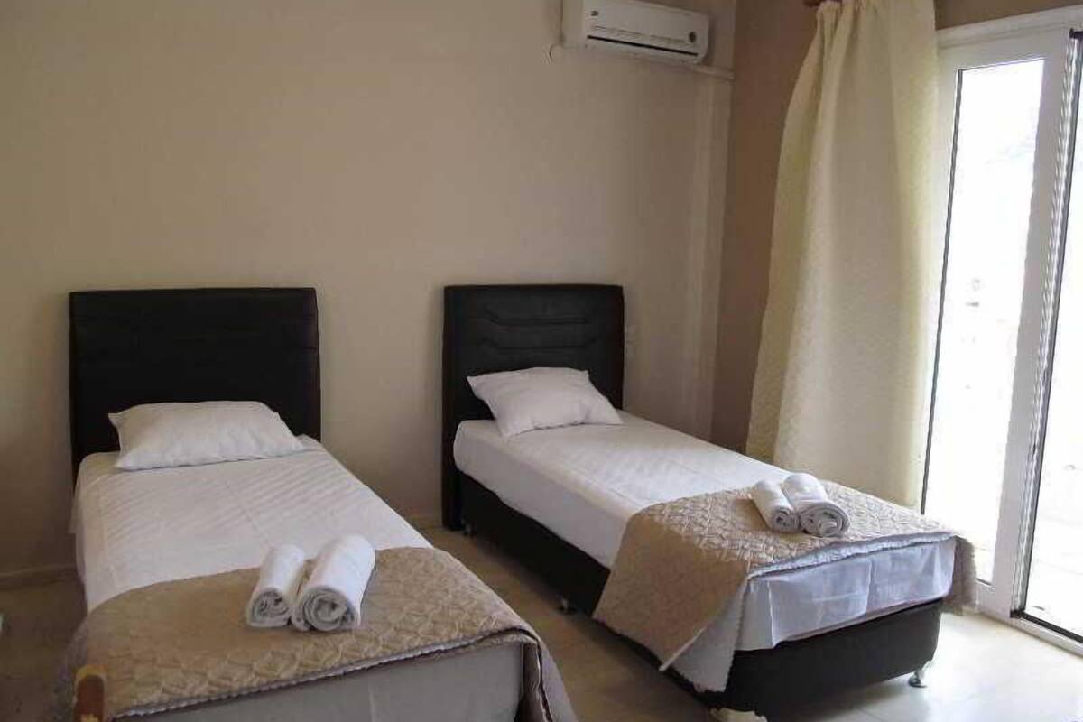 A hotel room with two single beds, a window with curtains, an air conditioning unit, and a view of the outside.