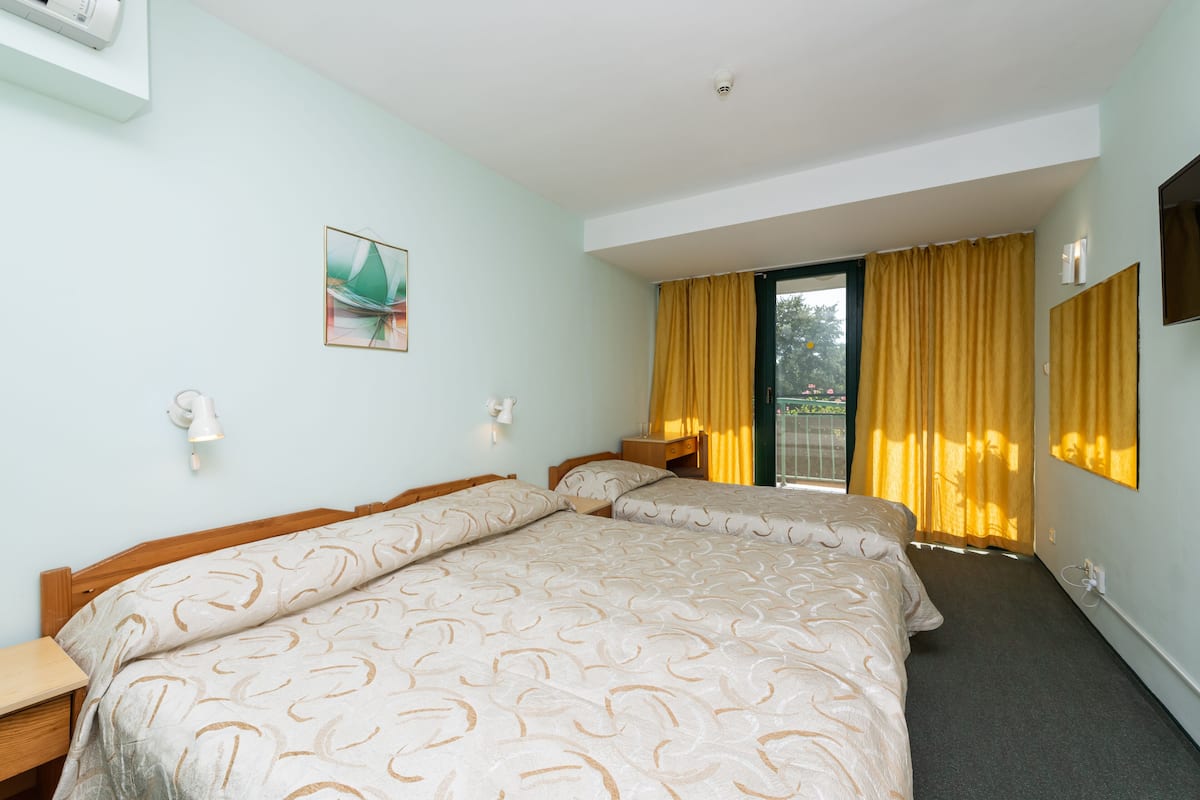 Habitación de hotel con dos camas, una ventana con cortinas amarillas, un televisor montado en la pared y un cuadro.