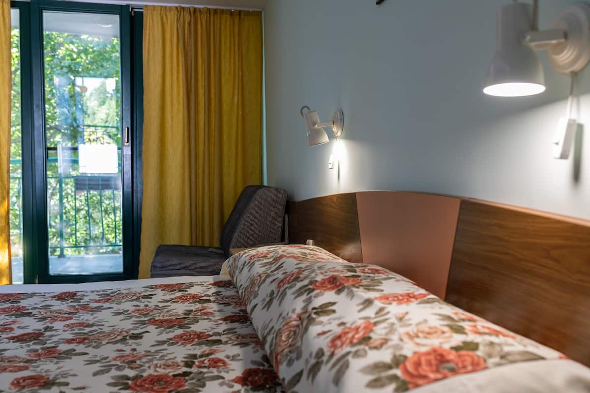 Habitación de hotel con cama, una silla, una mesita de noche, una lámpara y vistas al exterior.