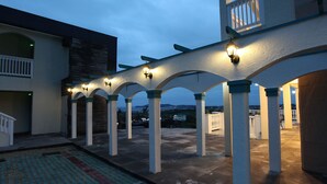 En udendørs overdækket terrasse med en række buer og søjler, udsigt over det omkringliggende landskab og en bygning med en balkon.