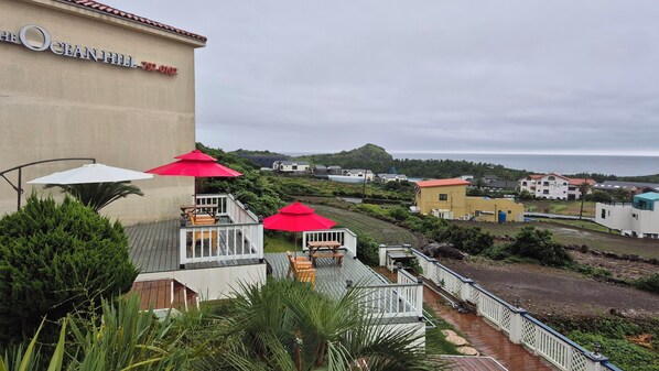 A coastal hotel with a deck area featuring outdoor seating and umbrellas, overlooking a beach and residential buildings. - The Ocean Hill (Seogwipo)