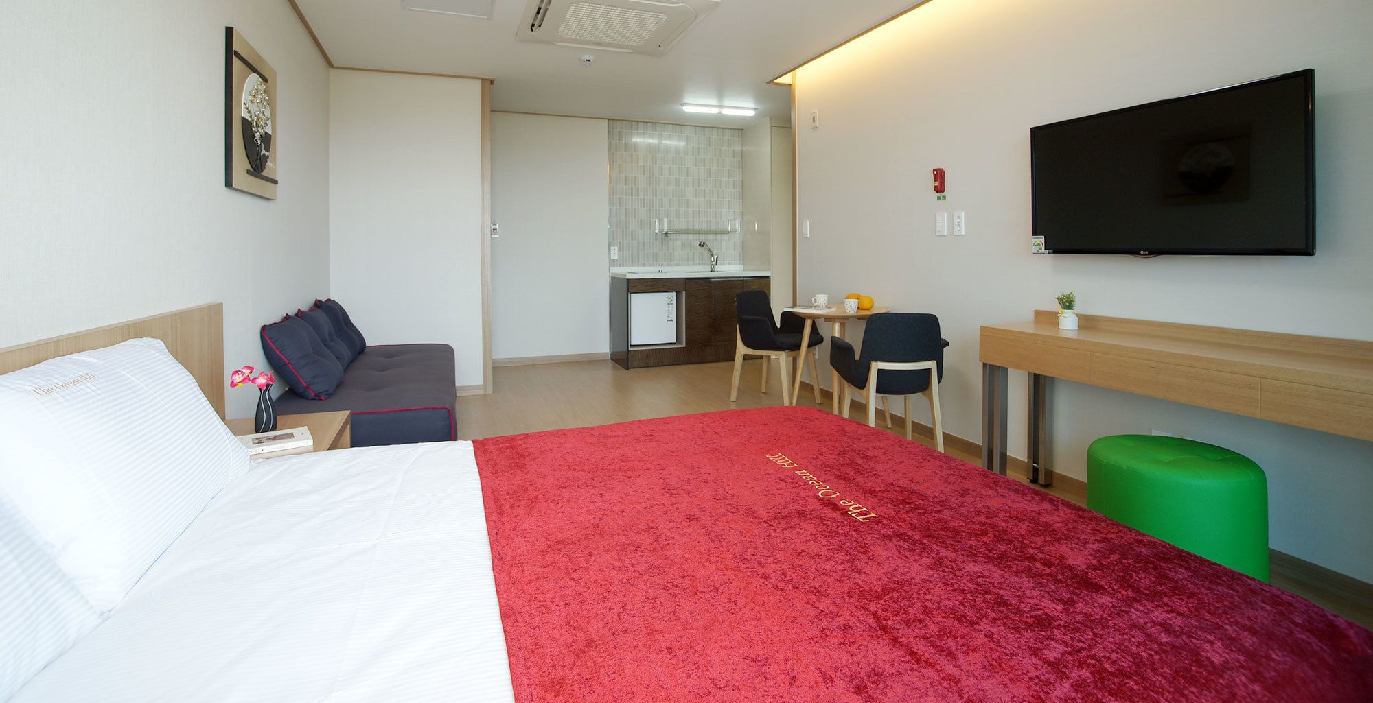 A hotel room with a bed, a desk, a chair, a small table with a cup and orange, and a green trash bin.