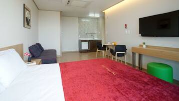 A hotel room with a bed, a desk, a chair, a small table with a cup and orange, and a green trash bin.