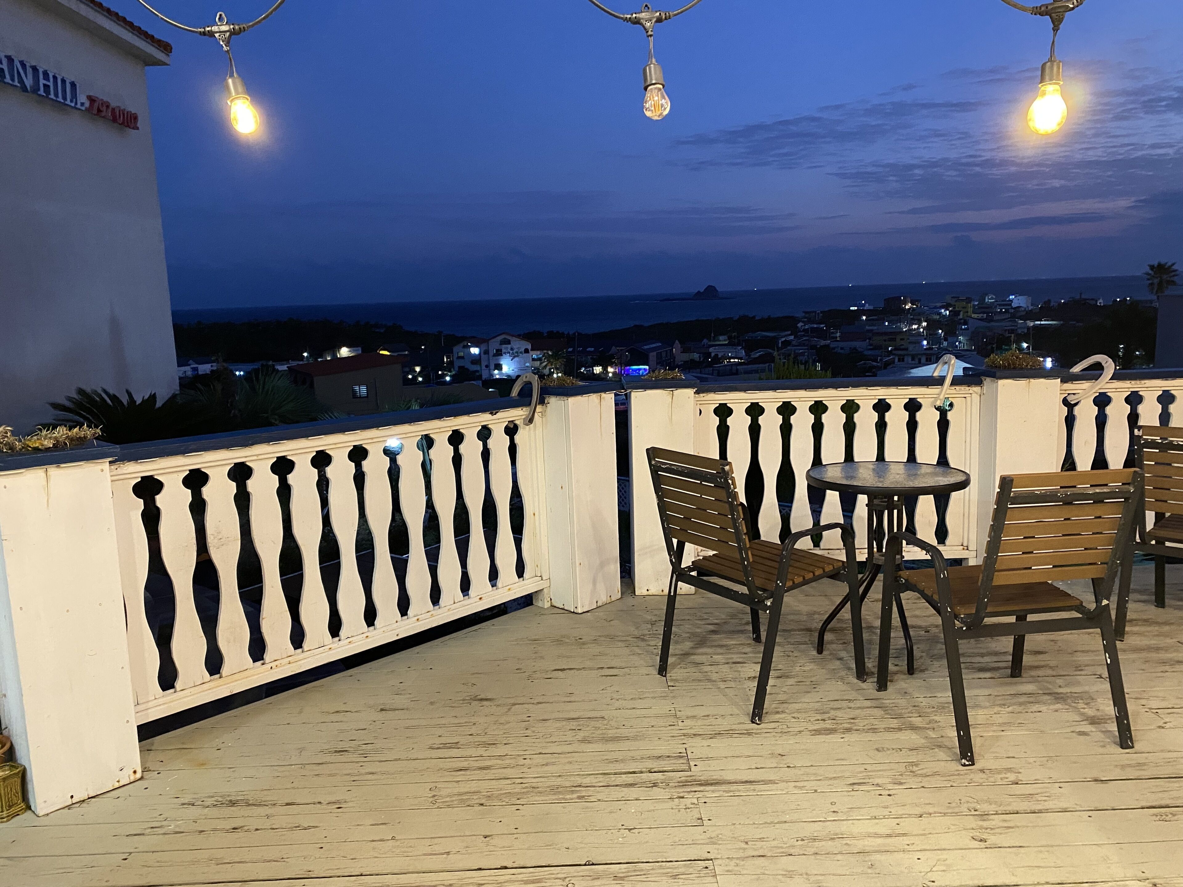 A balcony with a view of the city at dusk, featuring a table and chairs, a railing, and hanging lights.