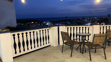 A balcony with a view of the city at dusk, featuring a table and chairs, a railing, and hanging lights.