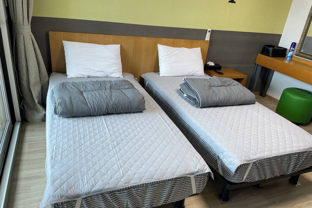 Une chambre d’hôtel avec deux lits, chacun doté d’un matelas et de coussins matelassés, d’une tête de lit en bois, d’une table de chevet avec une télécommande et d’une poubelle verte.