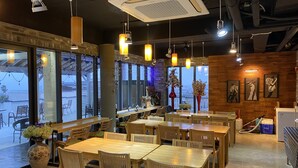 En restaurant med træborde og stole, store vinduer, hængende pendellamper og en murstensvæg.