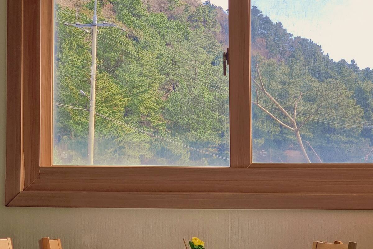 Une pièce avec une table dressée pour deux personnes, une fenêtre donnant sur un paysage montagneux et un vase contenant une seule fleur jaune sur la table.