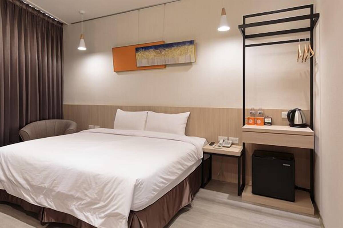 Une chambre d’hôtel avec un grand lit, un bureau avec une bouilloire, un téléphone et une petite poubelle.