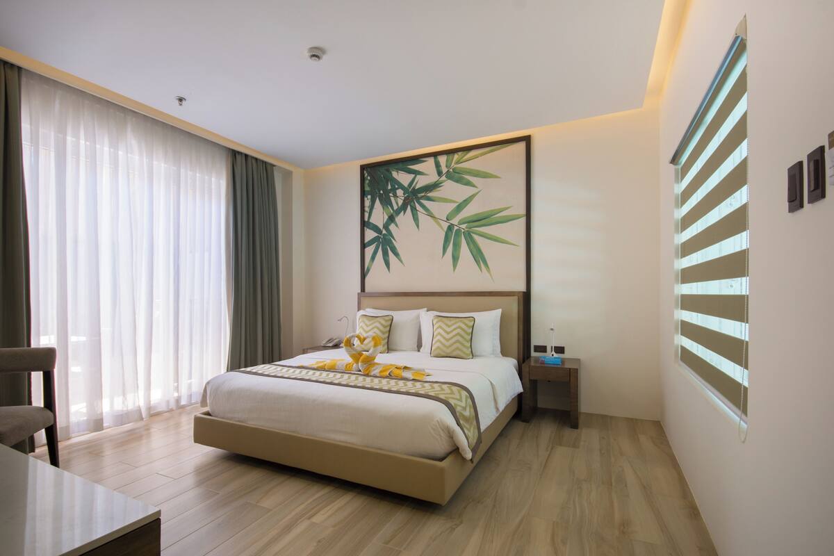 A hotel room with a large bed, a bedside table, a chair, and a window with blinds.