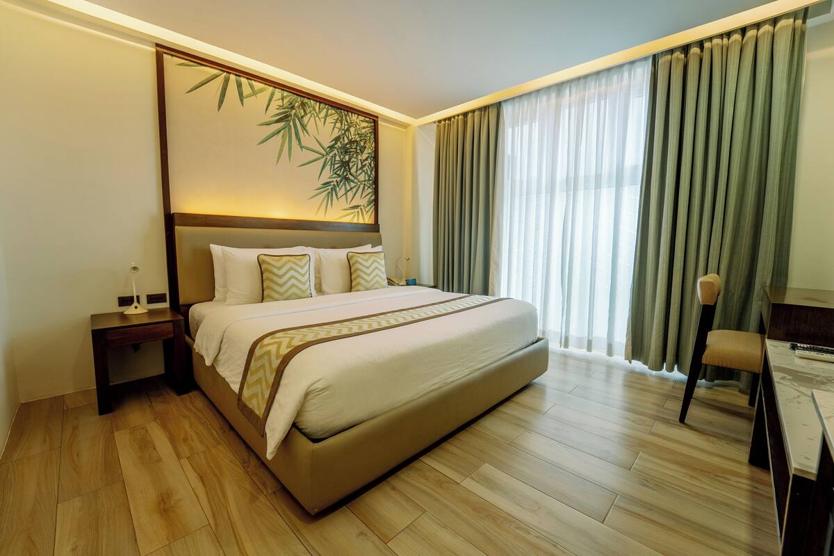 A hotel room with a large bed, a nightstand, a desk with a chair, and a window with curtains.