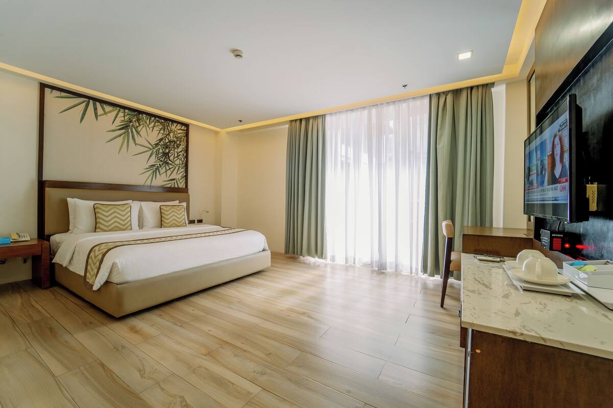 A hotel room with a large bed, a flat-screen TV, a wooden floor, and a large window with curtains.