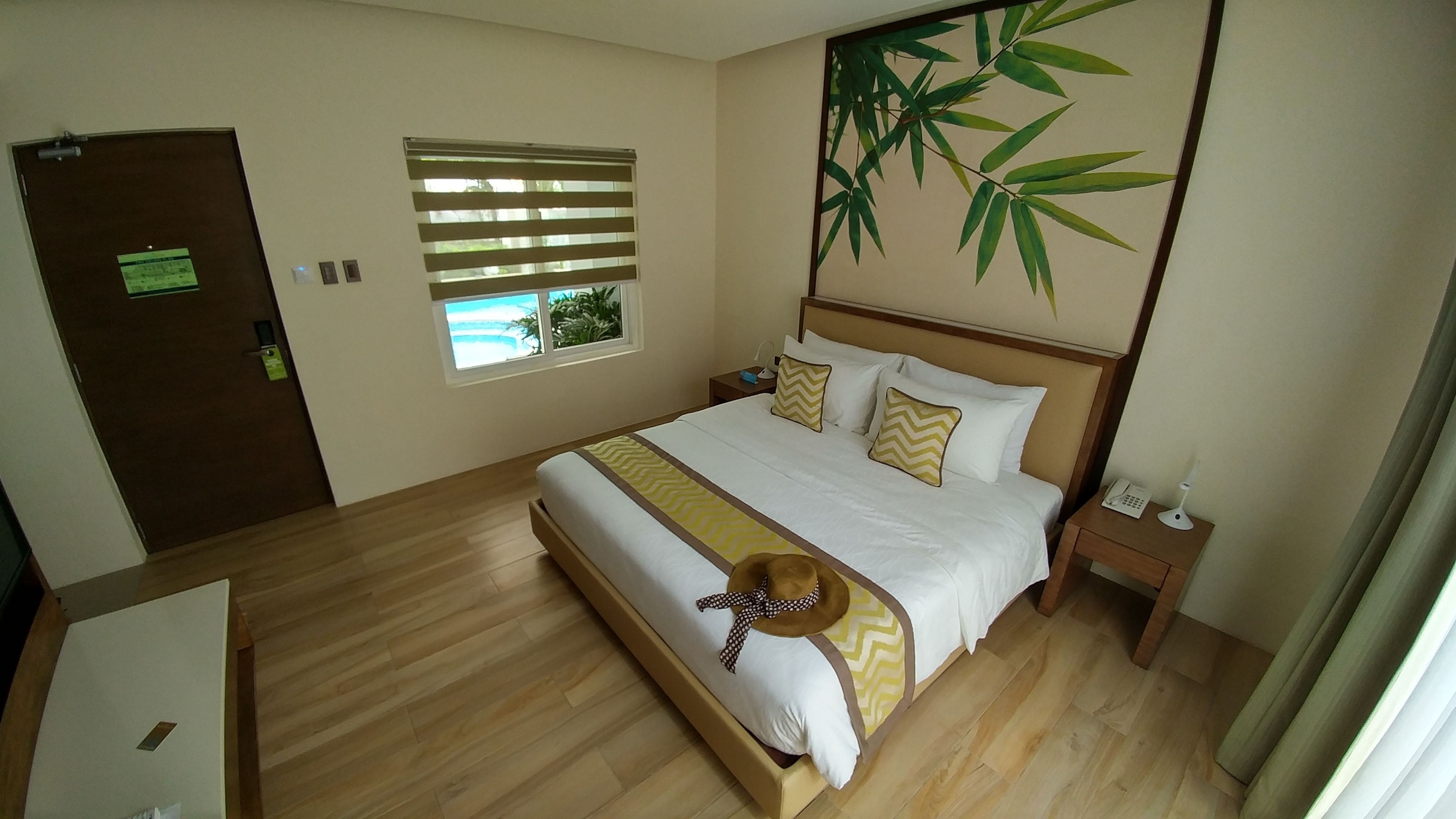 A hotel room with a bed, a nightstand, a window with blinds, a door, and a wall with a bamboo plant decoration.