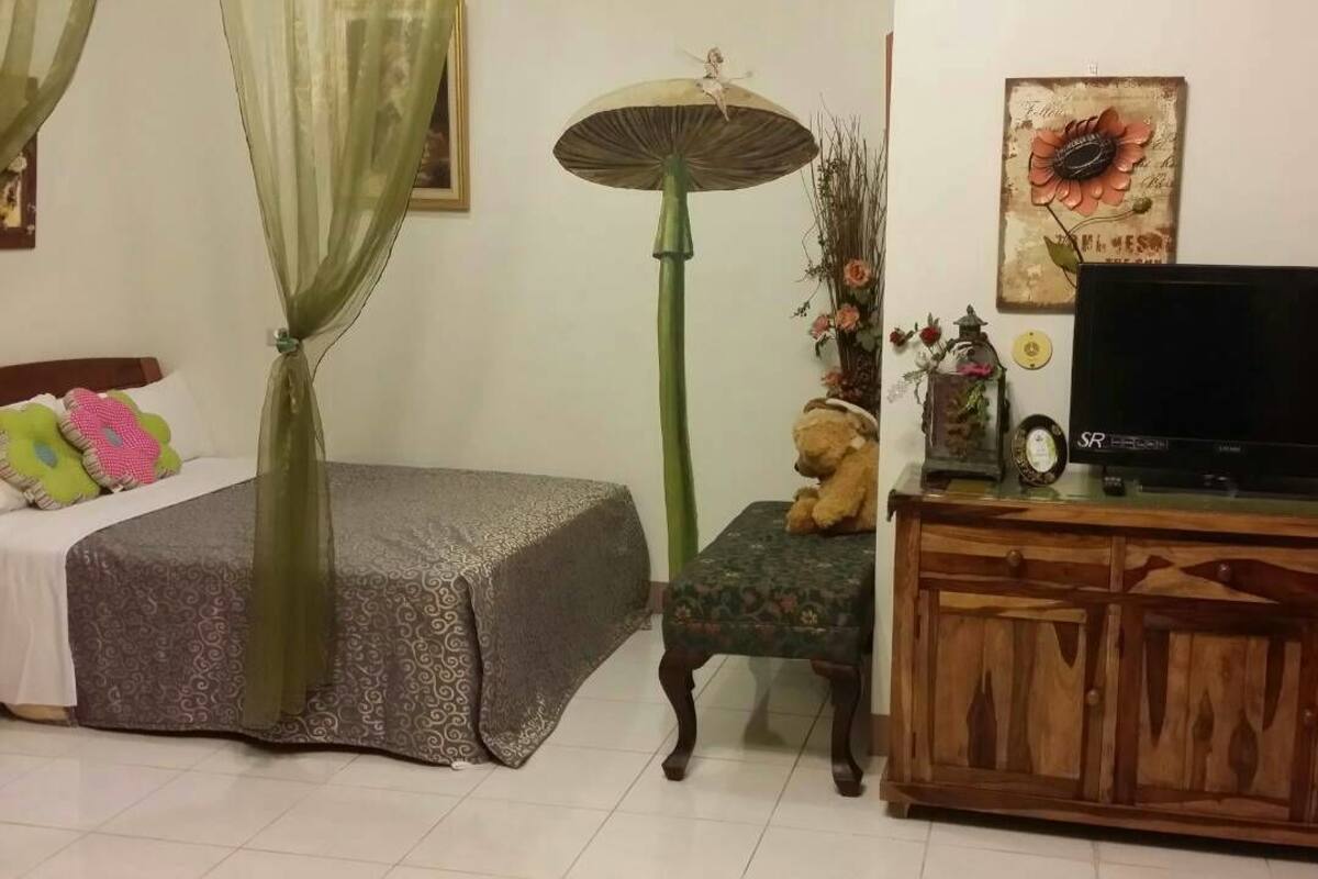 Una camera da letto con un letto, una lampada da terra verde, un televisore su un mobile in legno e un piccolo tavolino con oggetti decorativi.