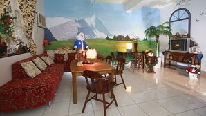 A cozy living room with a large sofa, a dining table, and chairs. There is a mural on the wall depicting a snowy mountain landscape.