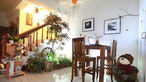 A cozy living room with a wooden staircase, a dining table, chairs, a wooden rocking chair, and various plants.