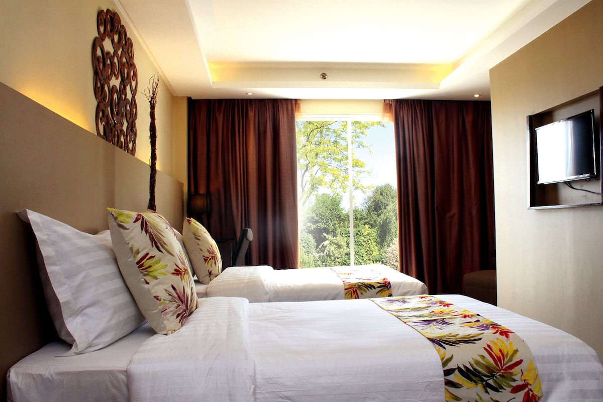 A hotel room with two beds, a flat-screen TV, a decorative wall piece, and a view of trees through the window.