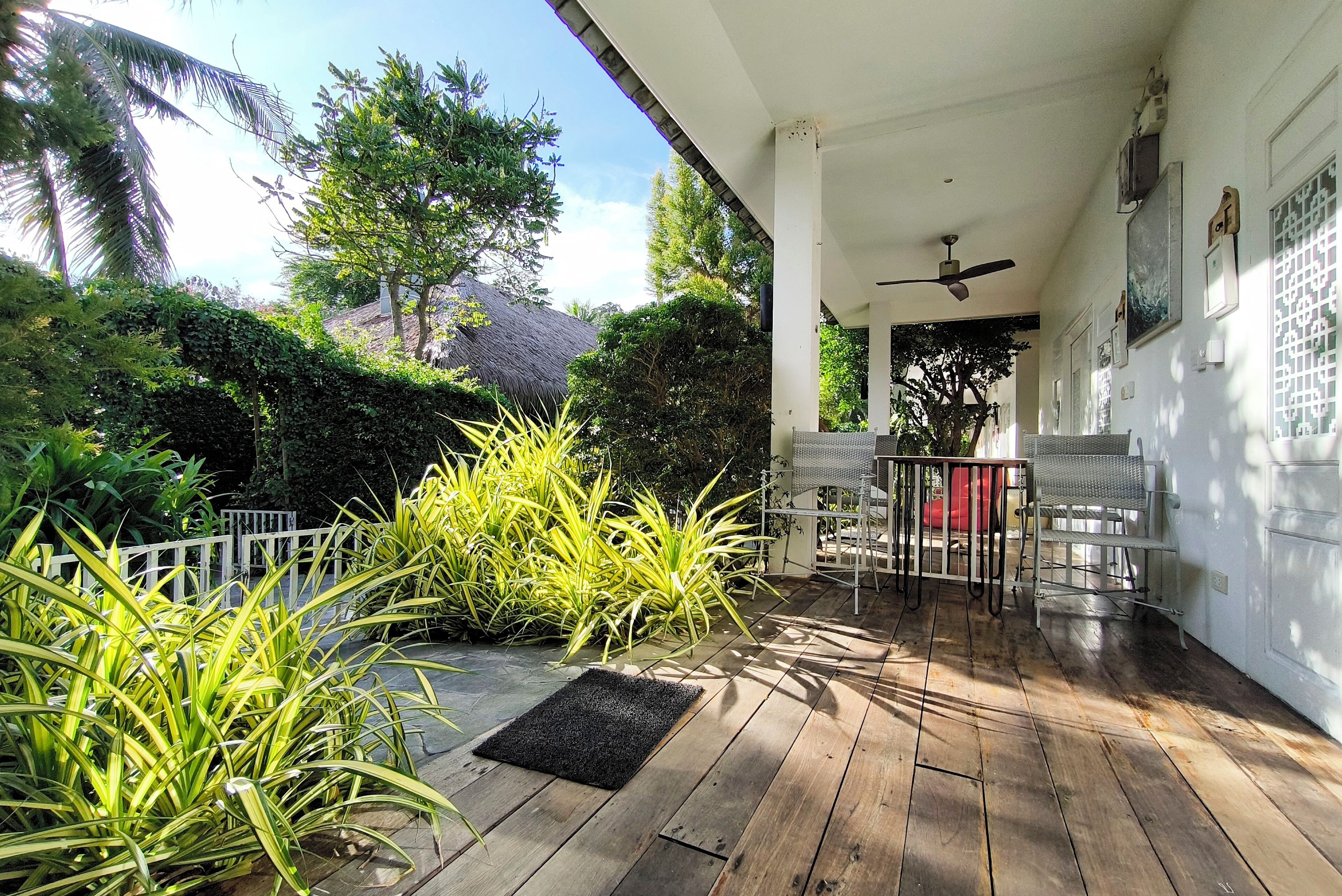 Una terraza de madera con ventilador de techo, mobiliario exterior y abundante vegetación.