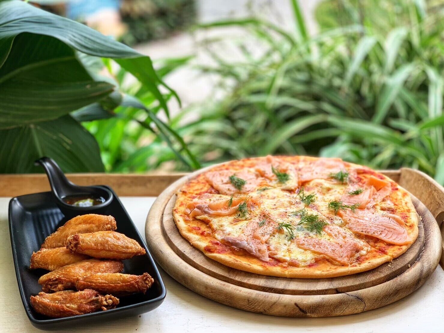 A pizza with salmon and herbs on a wooden board, accompanied by a black dish of fried chicken wings.