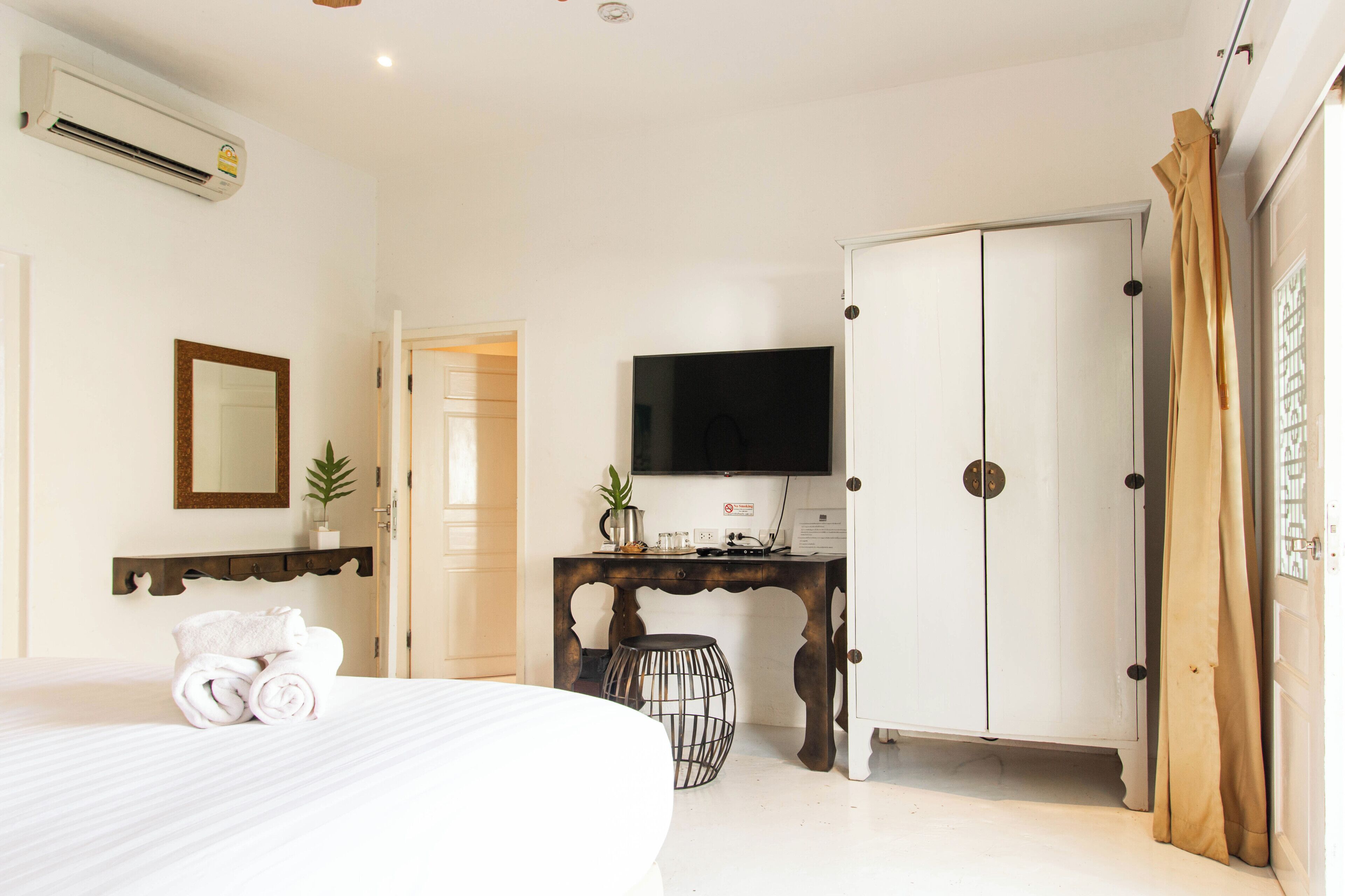 A hotel room with a bed, a TV, a wardrobe, a desk with a plant, and a mirror on the wall.