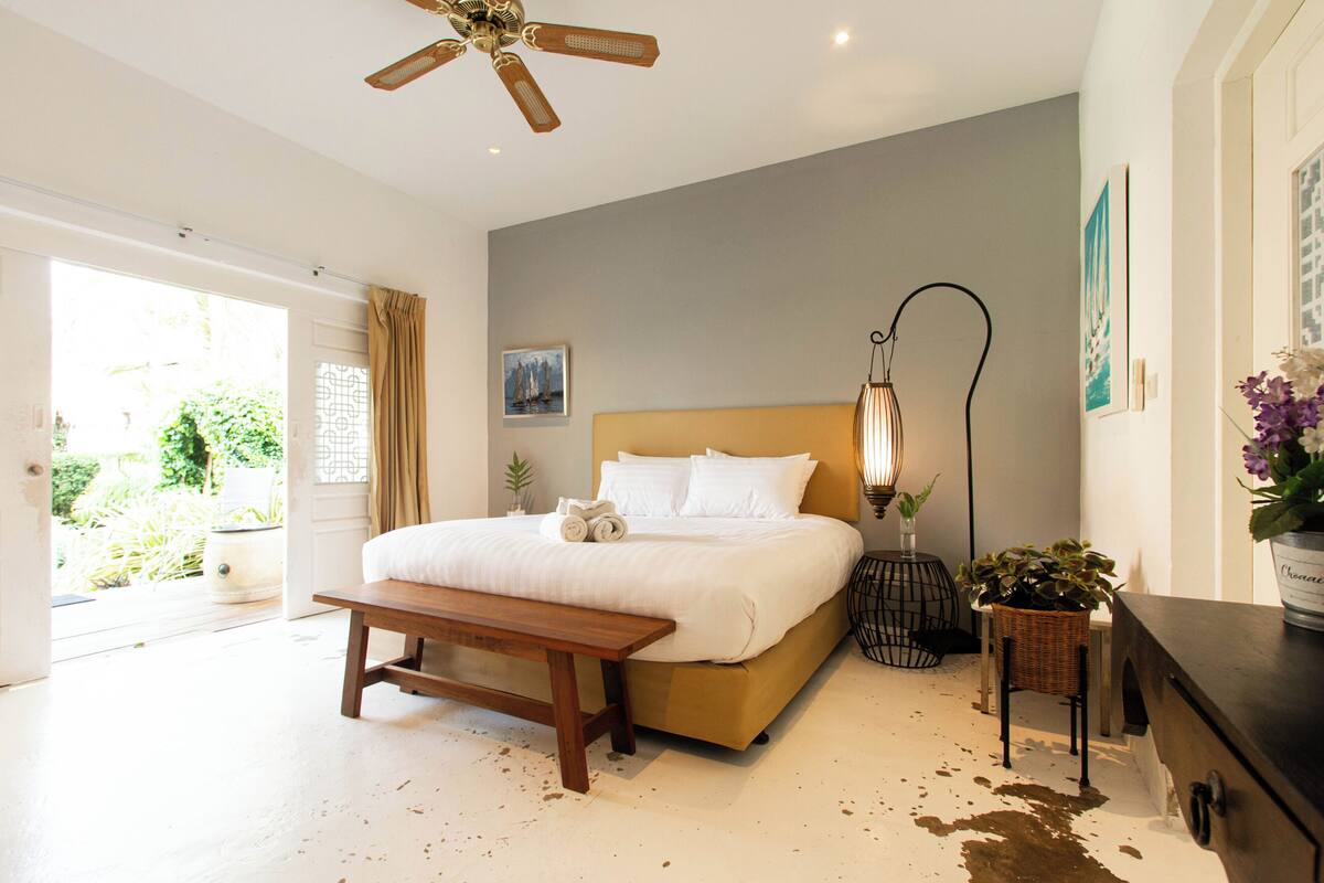 Ein Schlafzimmer mit einem Bett, einer Bank, einem Nachttisch, einer Stehlampe und einem Blumentopf.