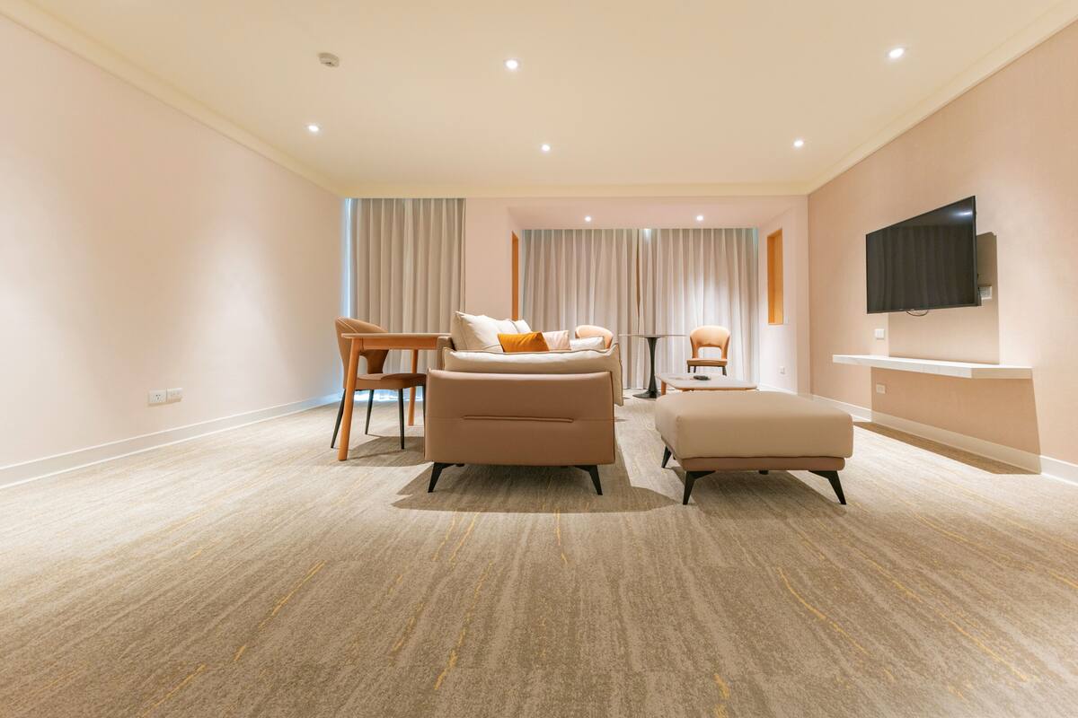 Ein modernes Hotelzimmer mit Sofa, Hocker und Sesseln, einem flachbildfernseher an der Wand und einem großen Fenster mit Vorhängen.