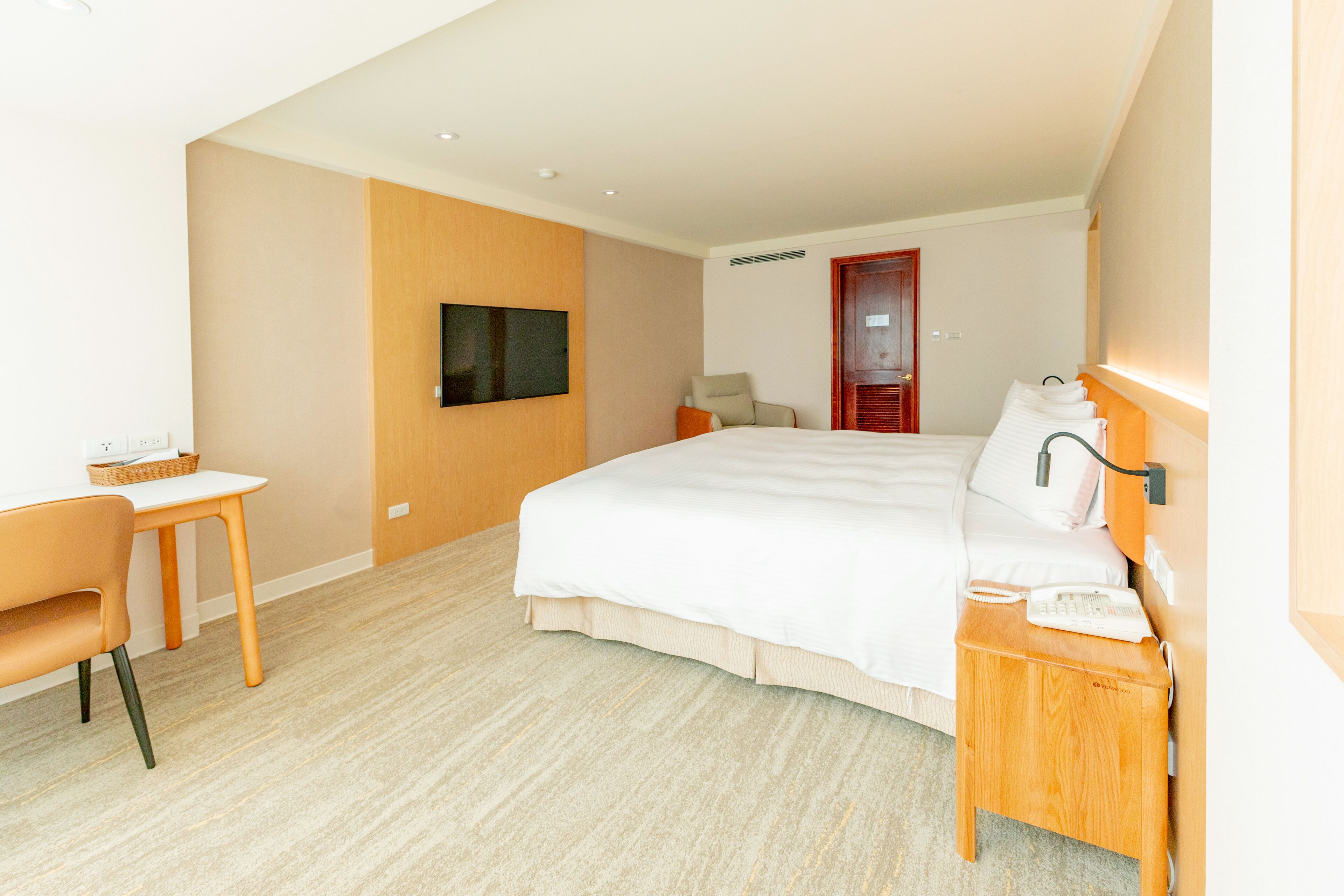 A hotel room with a large bed, a desk, a chair, a TV mounted on the wall, and a wooden nightstand with a phone.