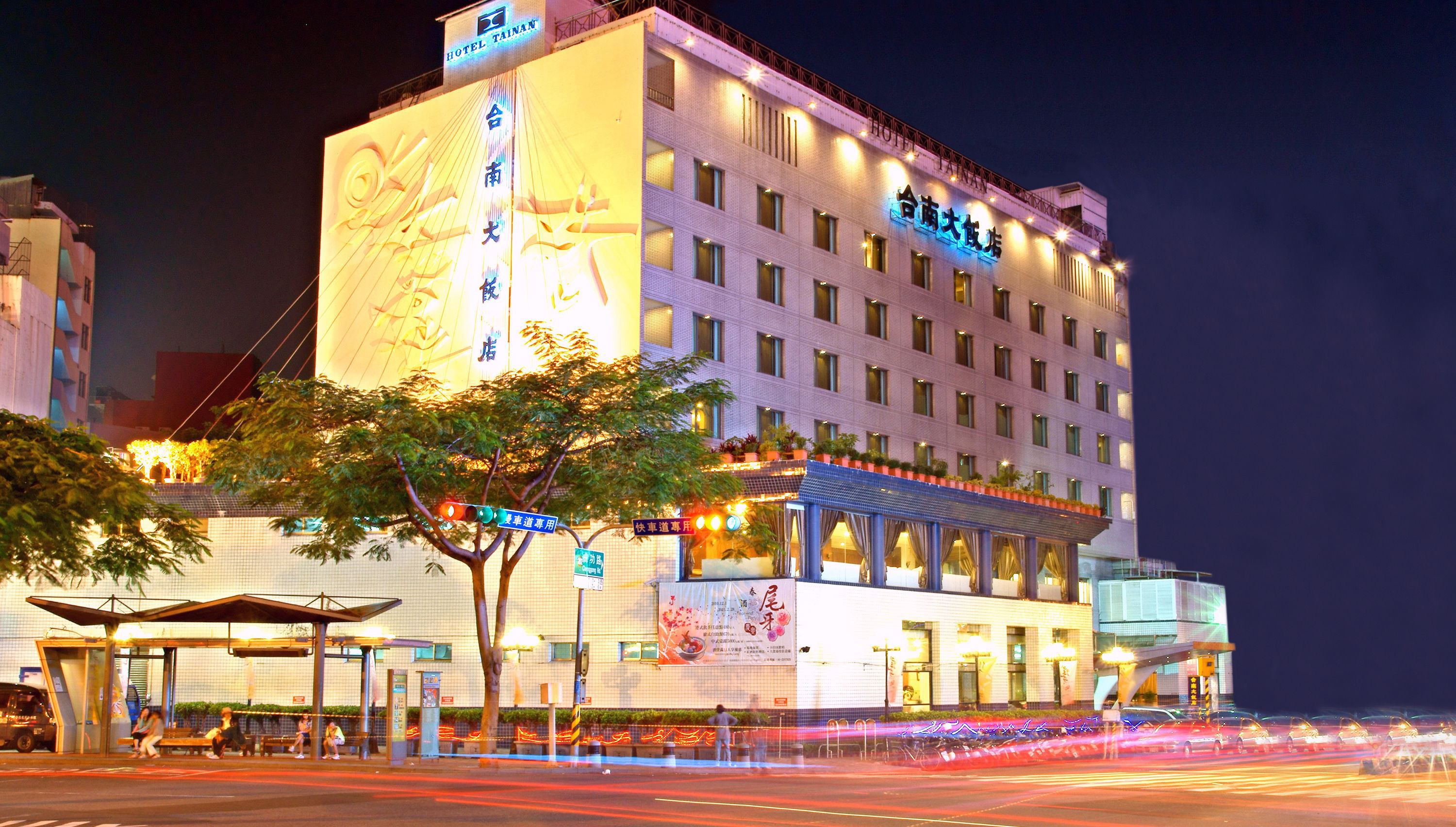 Un hotel de varios pisos con señalización iluminada, una entrada cubierta y una vista de la calle con estelas de luz.