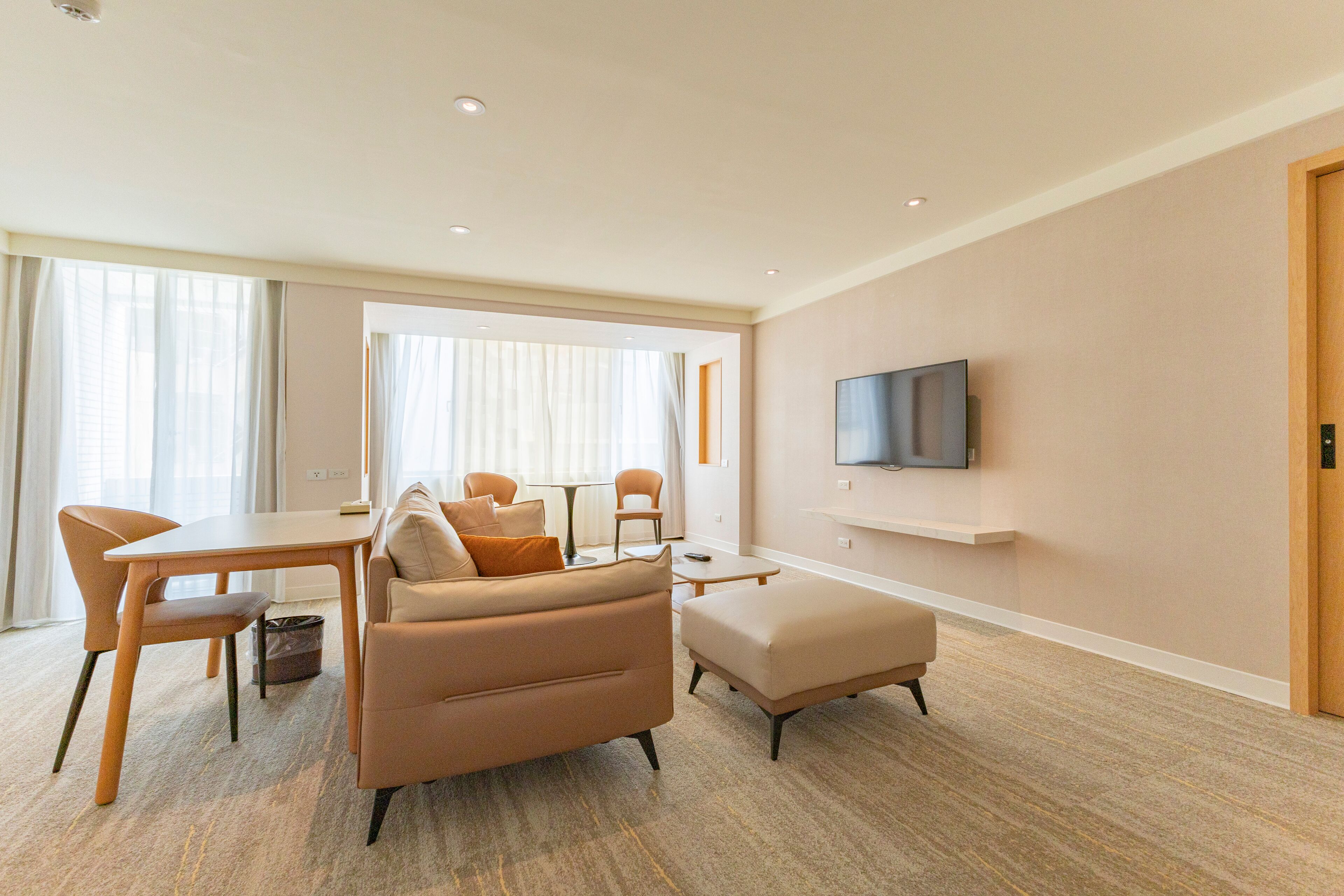 Ein modernes Hotelzimmer mit einer Couch, einem Esstisch, Stühlen, einem an der Wand montierten Fernseher und einem großen Fenster mit Vorhängen.