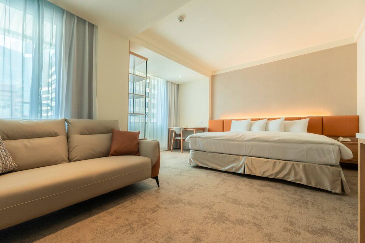 Ein Hotelzimmer mit einer Couch, einem Bett mit orangefarbener Kopfteil, einem kleinen Tisch und einem Fenster mit Vorhängen.