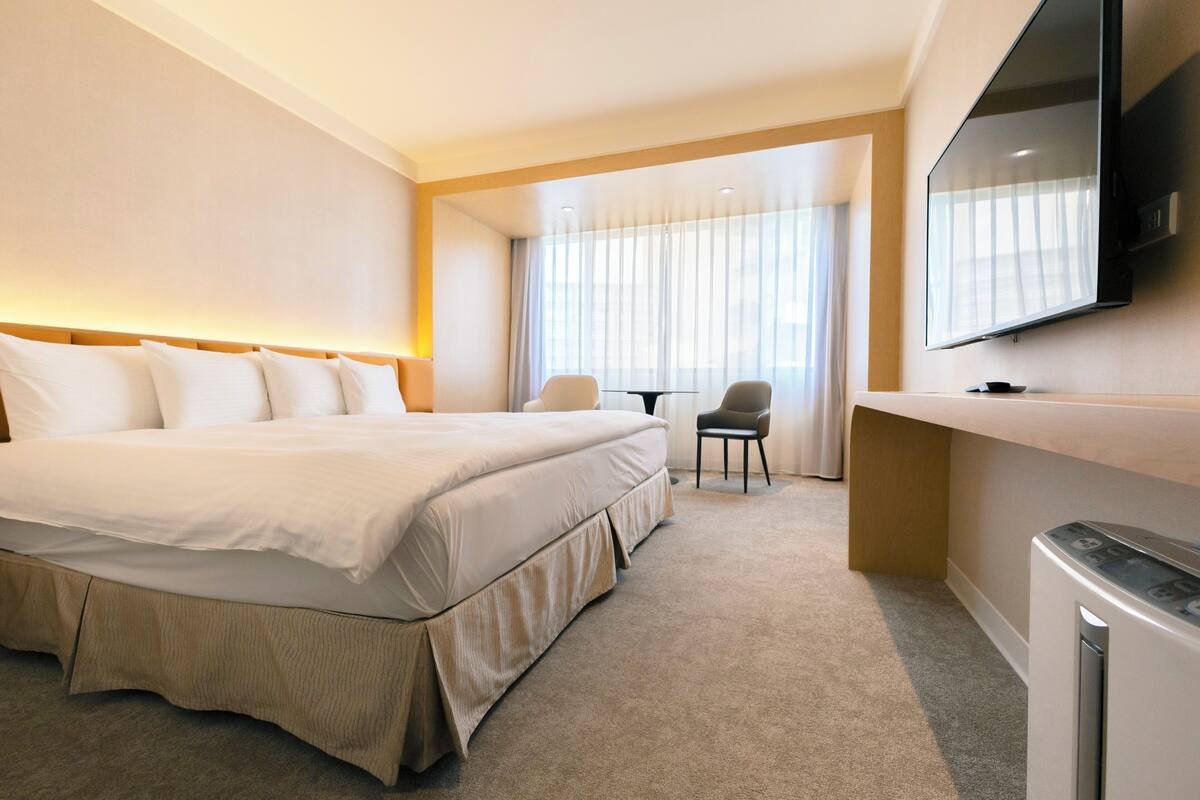 Ein Hotelzimmer mit einem großen Bett, einem an der Wand befestigten Fernseher, einem kleinen Tisch und einem Stuhl.