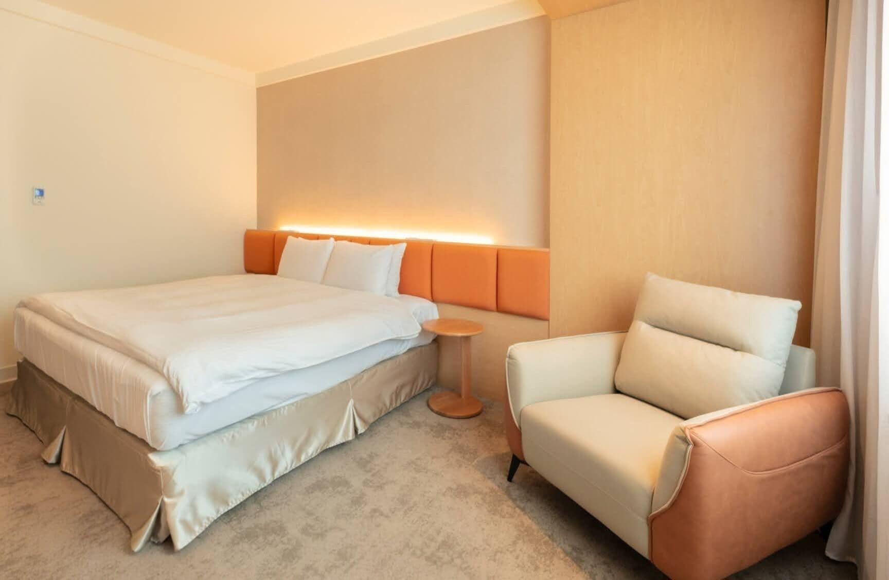 A hotel room with a bed, a chair, a small table, and a wall-mounted light.