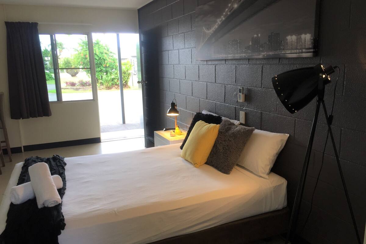 A modern hotel room with a large bed, a nightstand, a lamp, and a view of the outdoors.