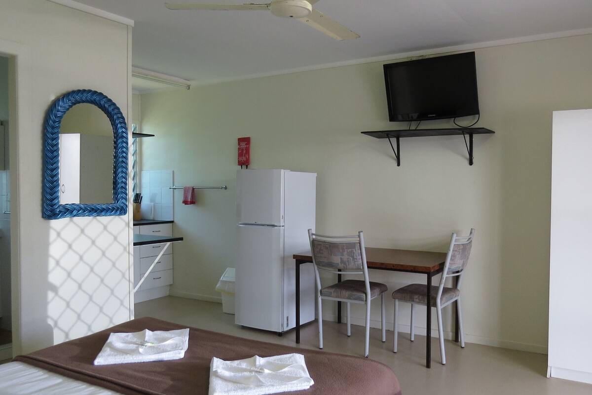 A compact room with a bed, a dining table with chairs, a small kitchen area with a refrigerator, a TV mounted on the wall, and a wardrobe.