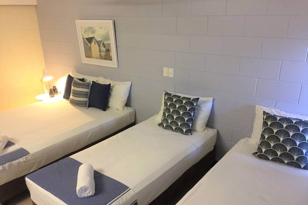 A hotel room with three single beds, each with a pillow and a rolled towel. There is a framed picture on the wall above the beds.