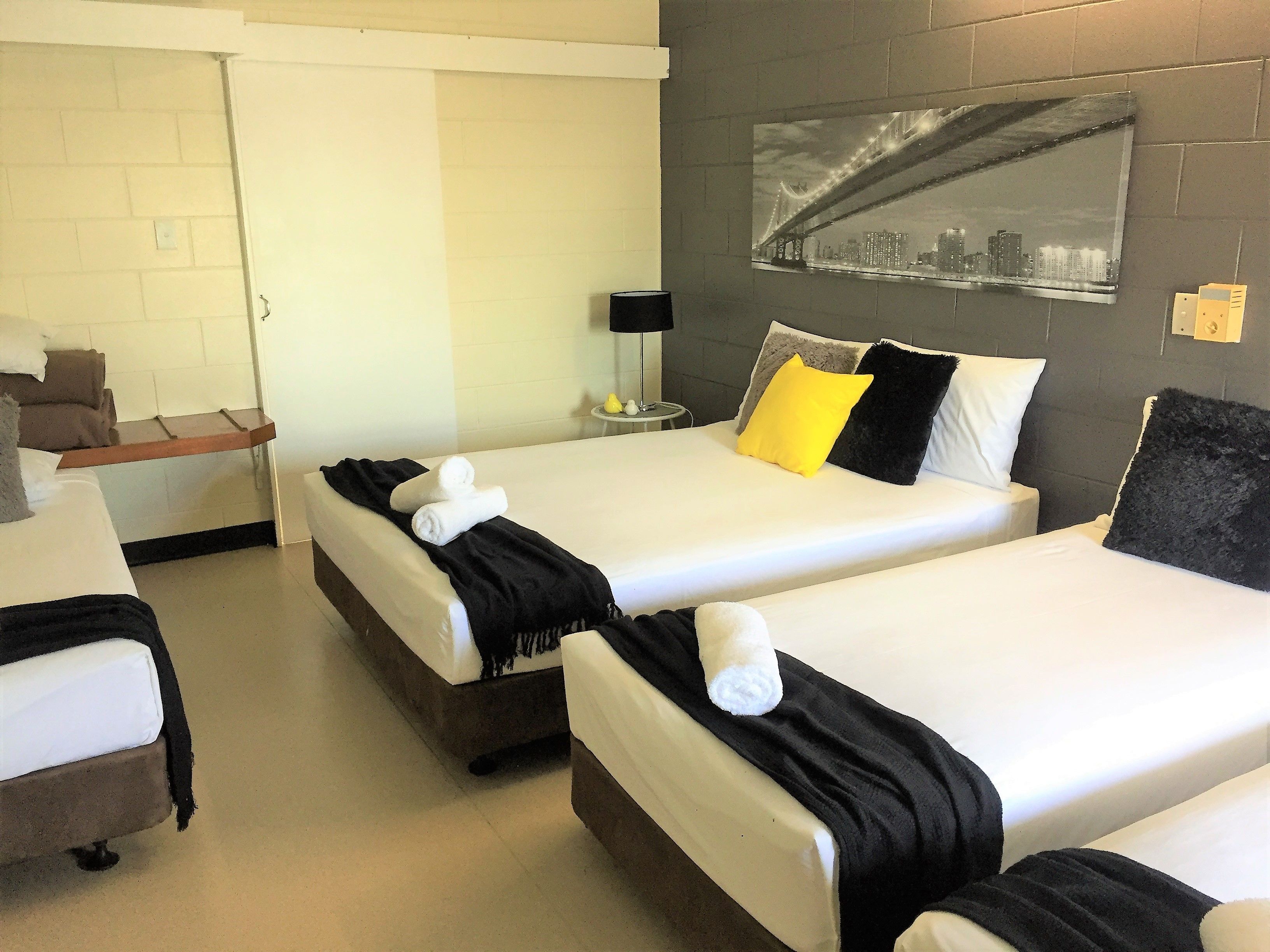 Habitación de hotel con dos camas, cada una con sábanas blancas y almohadas negras y amarillas. Hay una mesita con una lámpara y un espejo en la pared.
