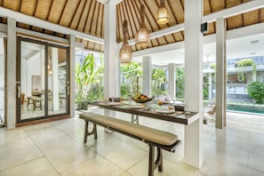 Uma sala de jantar espaçosa com uma grande mesa posta para refeição, rodeada por pilares brancos e um teto alto com vigas trançadas.