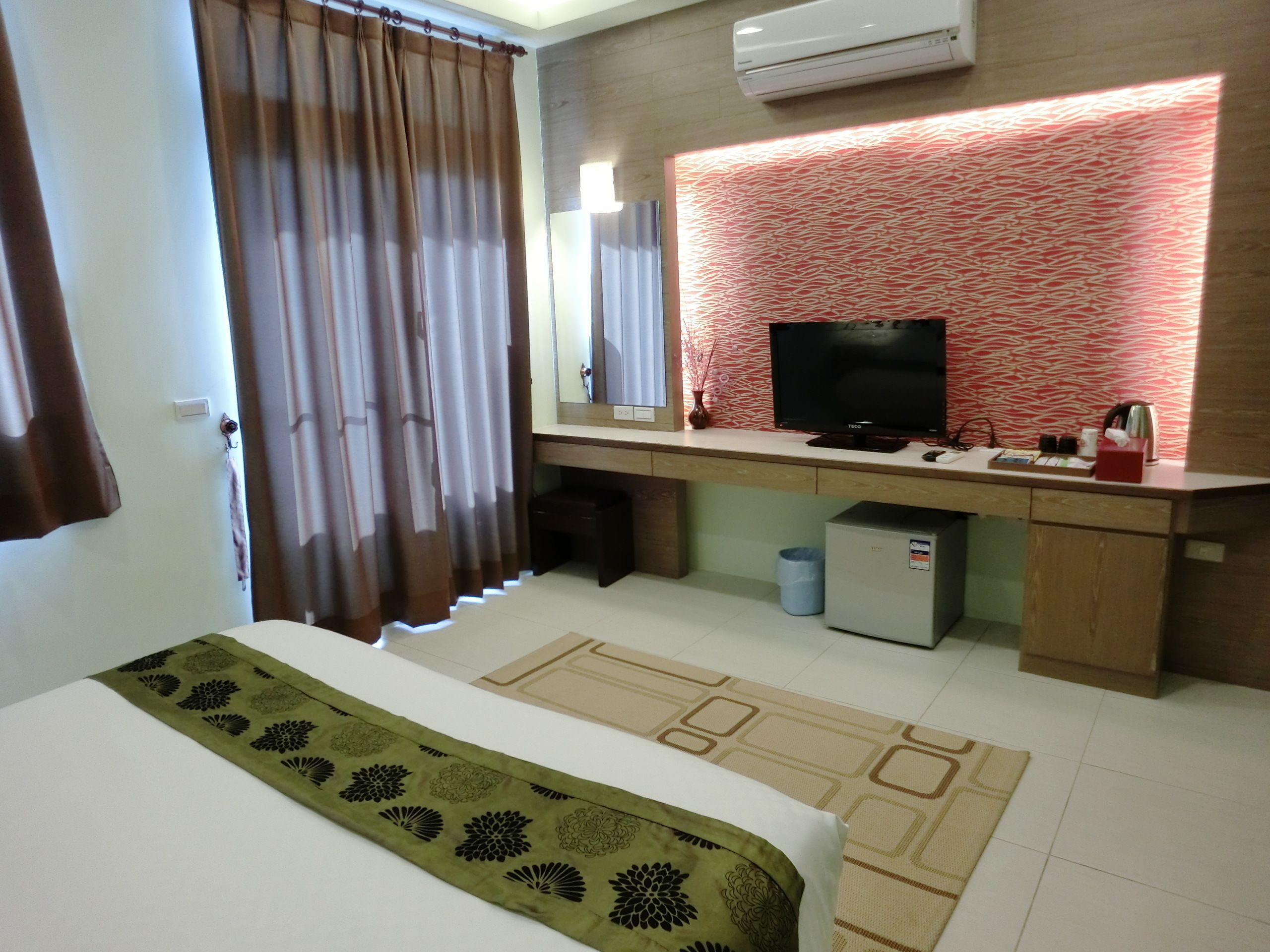 A hotel room with a bed, a desk with a TV, a small refrigerator, and a bathroom area.